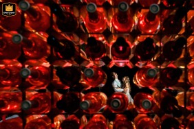   The wedding venue in Zuid Holland provides a creative backdrop as the bride and groom are artistically framed through red wine bottles in an imaginative composition.